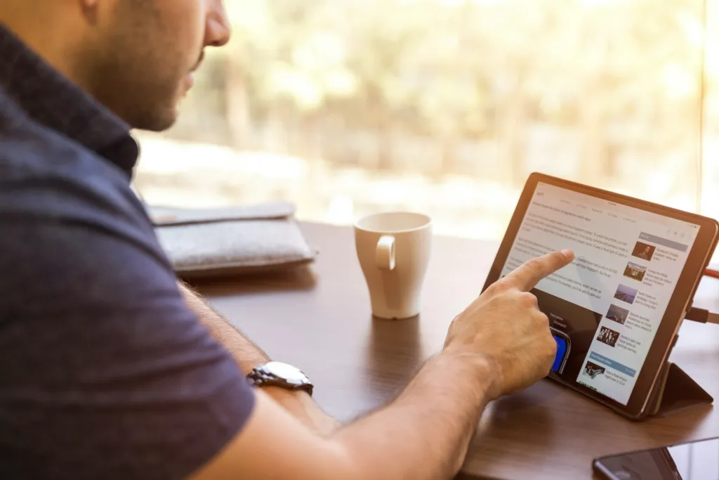 A man sits at a desk reviewing a content calendar on a tablet while holding a coffee cup, with a notebook and smartphone nearby, in natural window lighting.