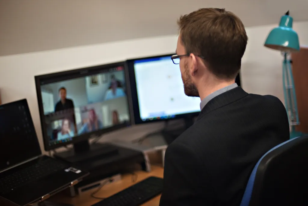 A man wearing glasses and a dark shirt sits at a desk facing a monitor displaying a video call with multiple participants. A laptop and a turquoise desk lamp are visible on the wooden desk. The man is engaged in what appears to be a live streaming or video conferencing session.