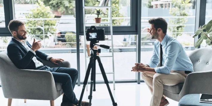 Two men sit in gray armchairs facing each other during a video recording session in a bright, modern office with large windows. A professional camera mounted on a tripod is positioned between them. Both men appear to be engaged in conversation, with one taking notes.