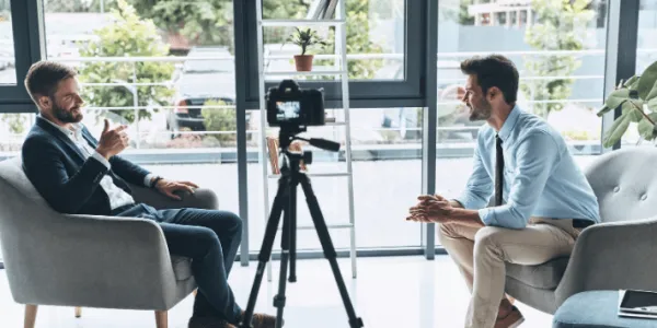 Two men sit in gray armchairs facing each other during a video recording session in a bright, modern office with large windows. A professional camera mounted on a tripod is positioned between them. Both men appear to be engaged in conversation, with one taking notes.