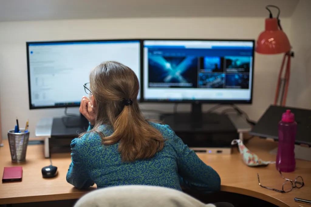 A woman with long brown hair and glasses sits at a wooden desk facing two monitors displaying video editing software and thumbnails. A red desk lamp, water bottle, and office supplies are visible on the desk beside her.