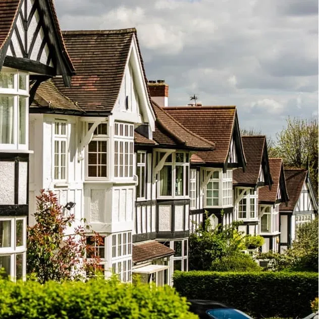 A row of Tudor-style houses with distinctive black and white timber framing, steeply pitched roofs, and large windows line a tree-lined street. The homes feature traditional architectural details including decorative half-timbering and terracotta roof tiles. Manicured hedges and vegetation frame the residential properties under a partly cloudy sky.