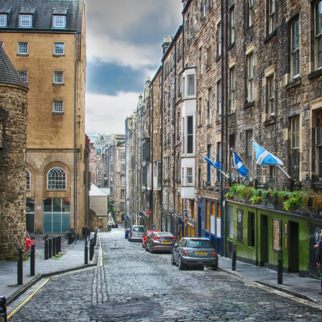 A narrow cobblestone street in Edinburgh's Old Town lined with historic stone buildings, some featuring yellow sandstone facades. Scottish flags hang from a storefront on the right, while parked cars and bollards line the street below a partly cloudy sky.
