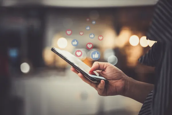 A hand holds a smartphone displaying floating social media icons including hearts and thumbs up symbols against a blurred urban background, representing digital engagement and online interaction for influencer marketing campaigns.