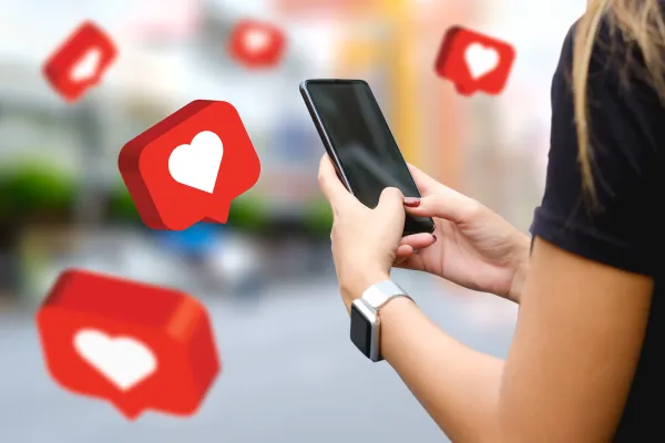 A person holds a smartphone while red heart-shaped social media notification icons float around them, representing likes and engagement on a social platform.