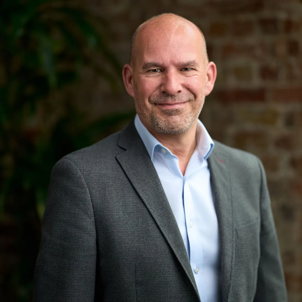 A bald man with a beard wearing a gray blazer and light blue shirt stands against a brick wall background with green foliage visible to the left.