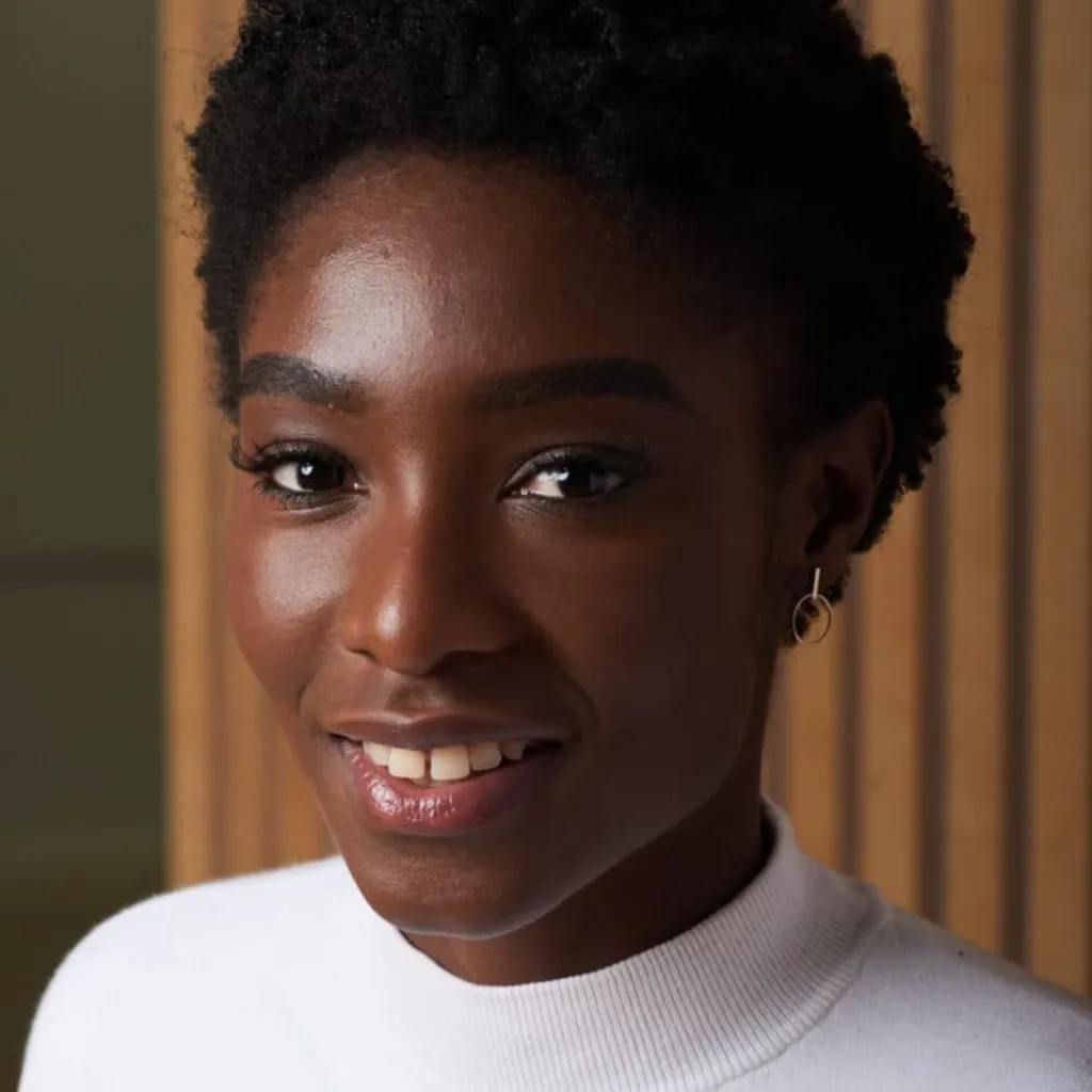 A woman with dark skin and short black hair, wearing a white ribbed turtleneck and small hoop earrings, smiles at the camera against a warm-toned interior background.
