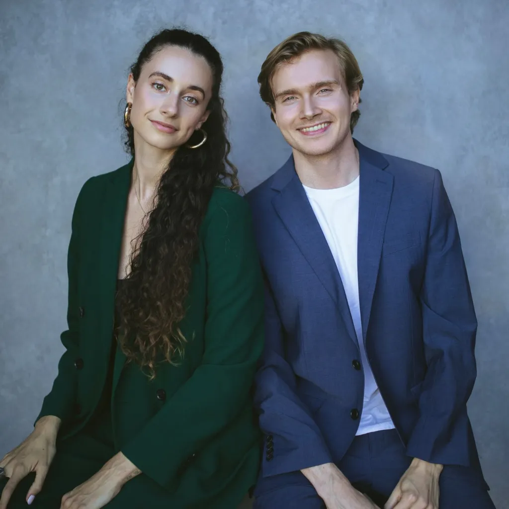 Two professionals pose together against a gray background. The person on the left wears a dark green blazer with hoop earrings and long wavy hair, while the person on the right wears a dark blue blazer over a white shirt with blonde hair.