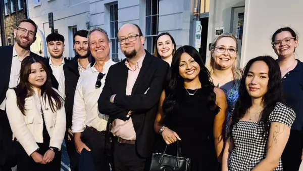 A diverse group of approximately 11 professionals stands together outside a modern building, smiling at the camera in a team photo. The group includes men and women wearing business casual and business formal attire, representing the team at Liz Male Consulting.