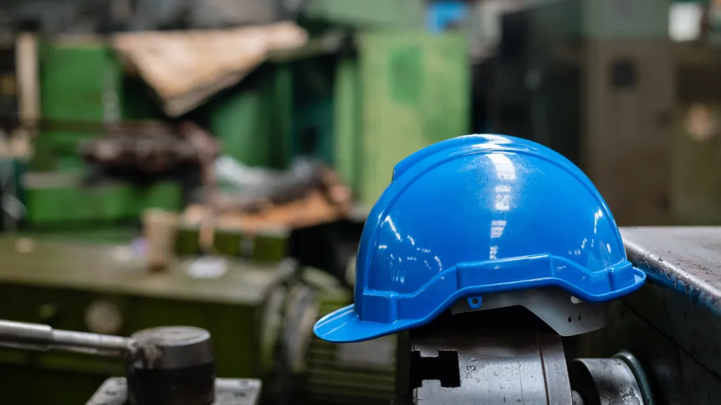 A blue hard hat rests on industrial machinery in a factory workshop, with blurred machinery and workbenches visible in the background, symbolizing construction and apprenticeship training environments.