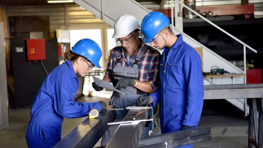 Three construction workers wearing hard hats and blue uniforms review blueprints together at a workbench in an industrial facility. The worker in the center, wearing a plaid shirt, points to details on the plans while the two colleagues in blue jackets observe. This represents collaborative learning and mentorship in construction apprenticeship training.