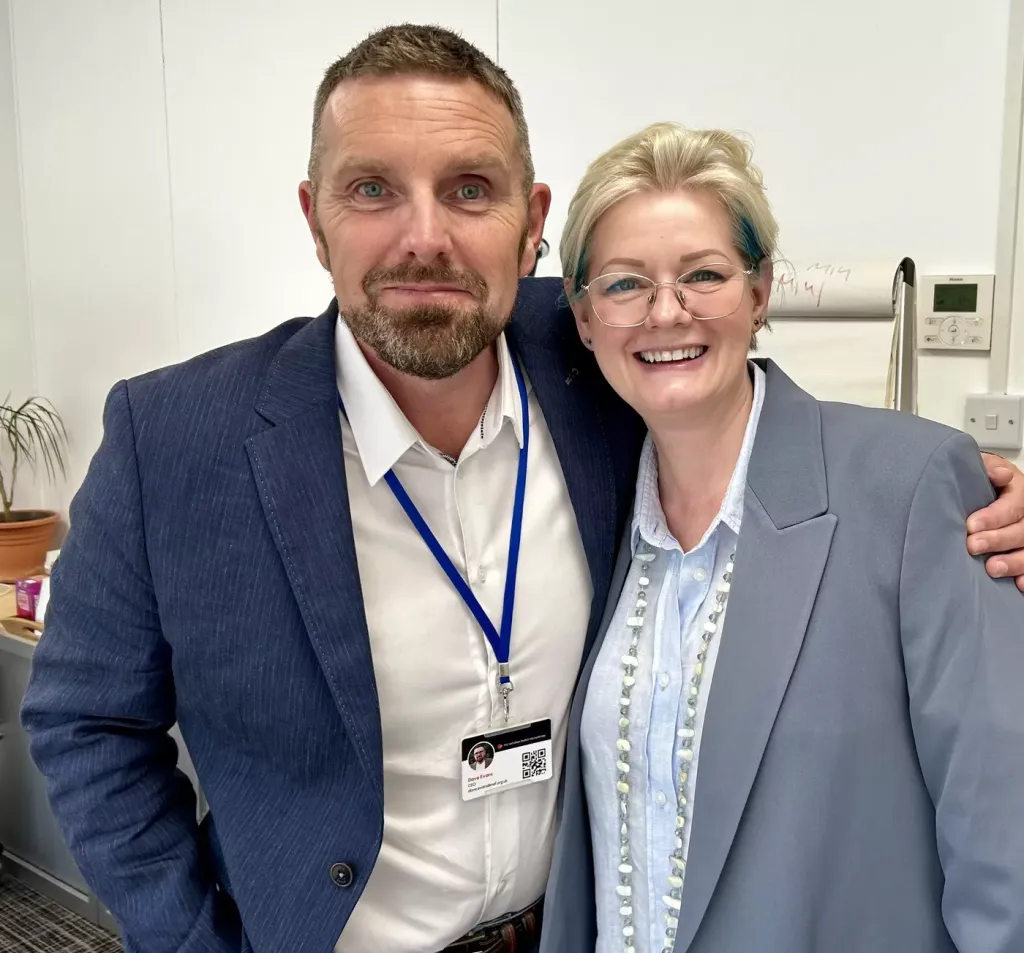 Two professionals pose together for a photo in an office setting. The man on the left wears a dark blue blazer with a white shirt and blue lanyard badge, while the woman on the right wears a light blue blazer over a white blouse with glasses. Both are smiling at the camera.