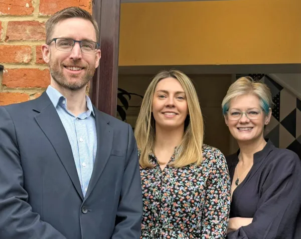 Three professionals stand together smiling at the camera in front a brick wall and gold-colored panel. A man in a navy blazer and light blue shirt stands on the left, a woman in a colorful floral dress in the center, and a woman in a dark blazer on the right.