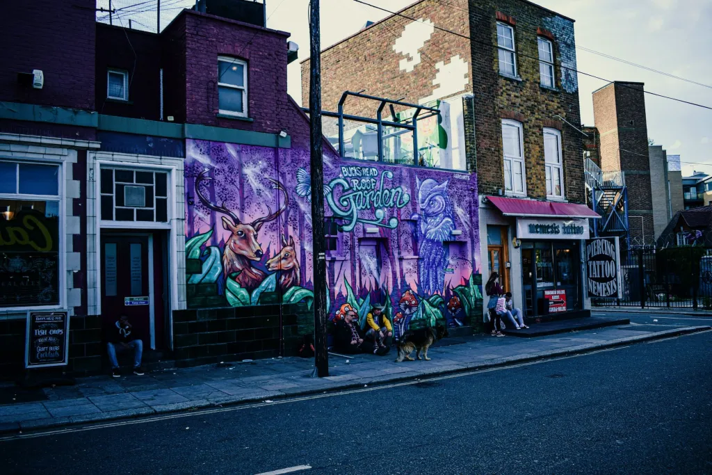 A vibrant purple and green street mural covers the side of a brick building on an urban street corner, with ornate lettering and artistic figures, while neighboring buildings and a street lamp frame the scene.