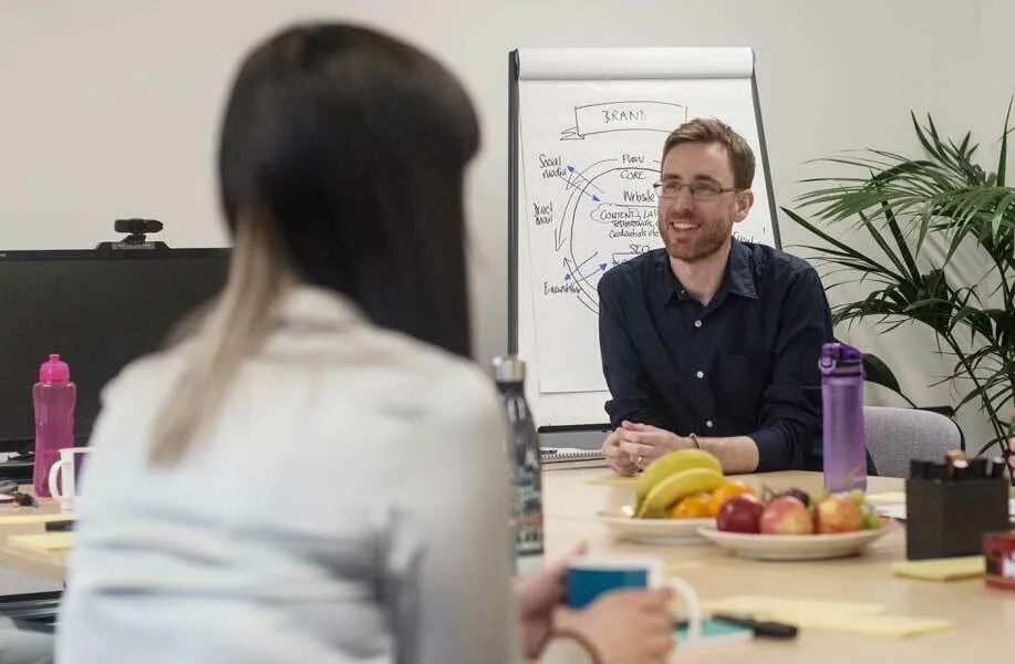 A man wearing glasses and a dark blue shirt sits at a desk across from a woman with long dark hair, with a whiteboard diagram visible behind him and fresh fruit and water bottles on the table between them.