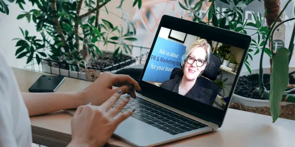 A person's hands gesture toward a laptop screen displaying a video call with a woman wearing glasses, with potted plants visible in the background of both the physical workspace and the video feed.