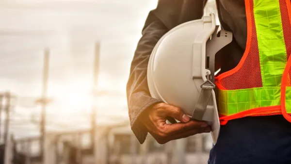 A construction worker in a high-visibility vest holds a white hard hat while standing at a job site during golden hour, with industrial structures visible in the blurred background.
