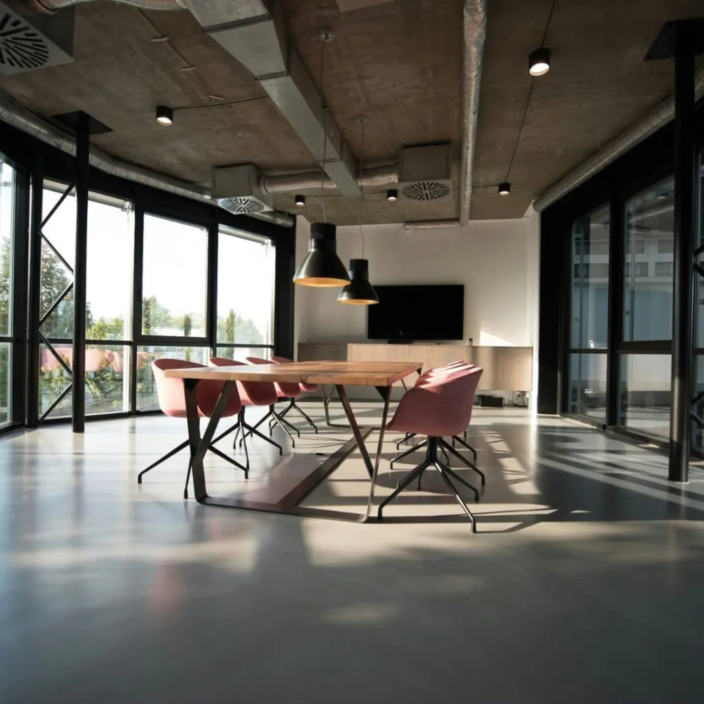 A modern office space with polished concrete floors, industrial ceiling, and large windows on either side. A wooden conference table with burgundy chairs sits in the center, with black pendant lights hanging above. Glass partition walls and a TV screen are visible in the background.