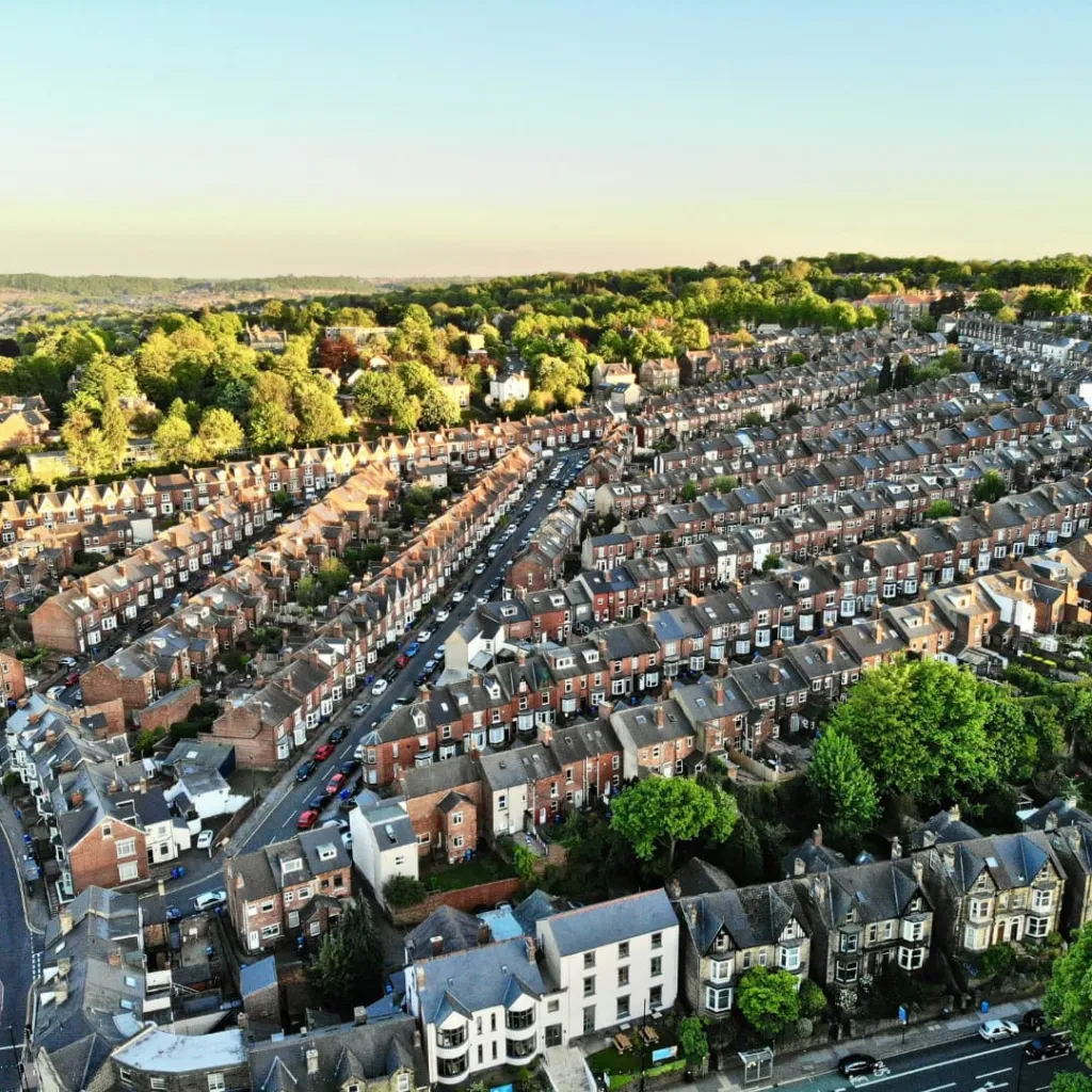 Aerial view of a dense suburban residential neighborhood with rows of brick townhouses and terraced homes arranged along winding streets, with green trees and parks visible in the background under clear sky.