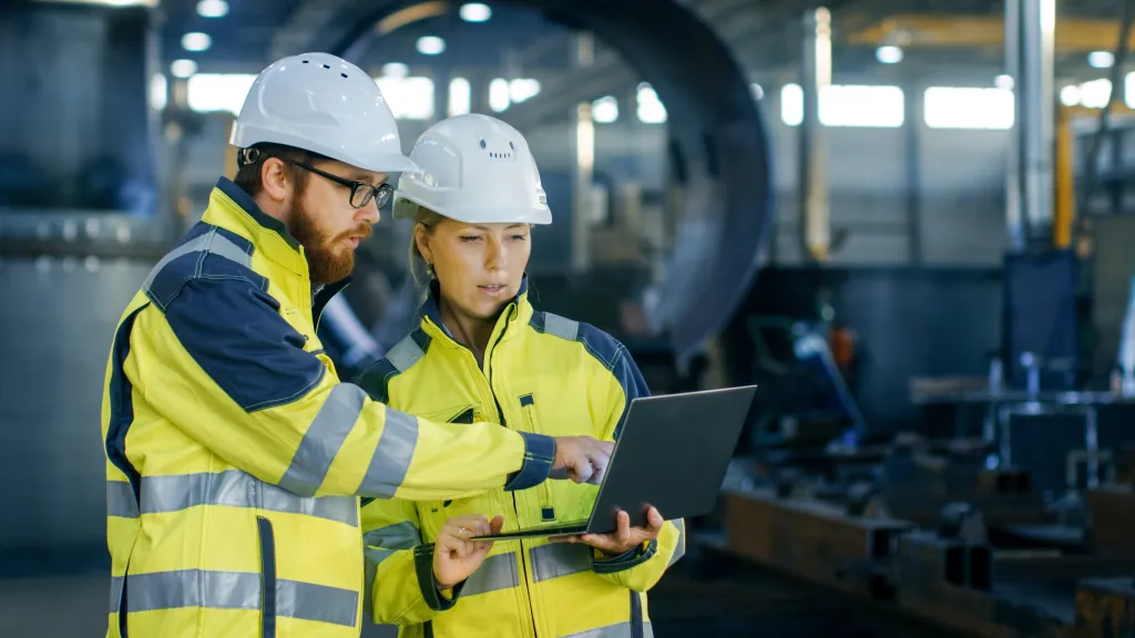 Two construction workers wearing white hard hats, yellow high-visibility vests, and safety glasses review a laptop together on an industrial job site. They appear to be discussing project details or safety protocols in a large facility with machinery and equipment visible in the background.