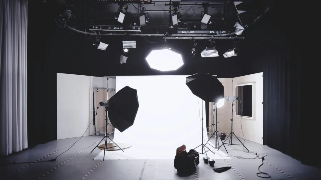 A professional photography studio with white backdrop and black walls, equipped with multiple studio lights on stands and overhead rigging, featuring softbox diffusers and professional lighting equipment arranged for product or portrait photography.