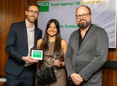 Three professionals stand together holding a PRCA award at an indoor event, with a banner visible in the background mentioning 