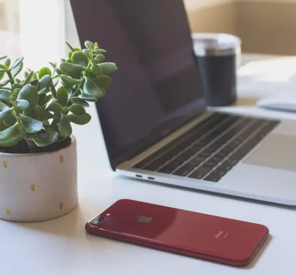 A red iPhone lies on a white desk next to a potted succulent plant, with a laptop and glass of water visible in the blurred background, representing a modern remote work setup.