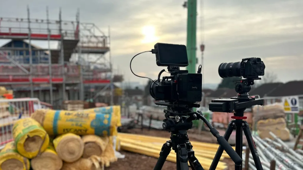 Two professional cameras mounted on tripods positioned at a construction site, with scaffolding, building materials, and yellow and blue storage containers visible in the background under an overcast sky.