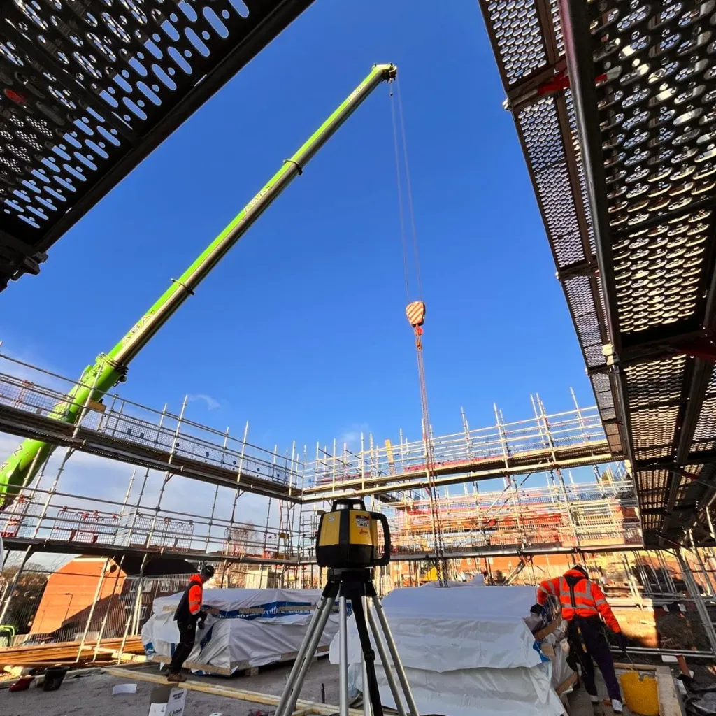 Construction workers in high-visibility vests operate surveying equipment on a steel-frame building site under clear blue sky, with metal scaffolding and safety netting visible overhead.