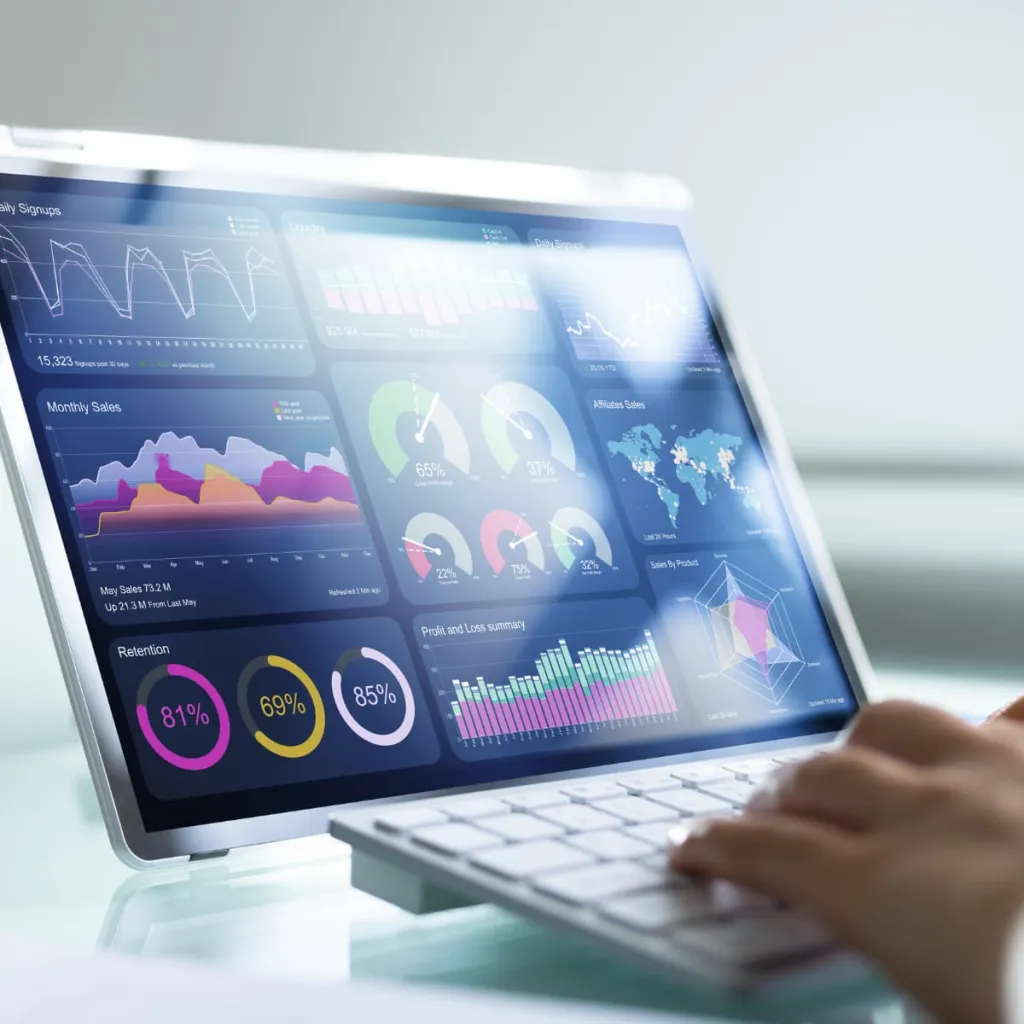 A person's hand operates a tablet displaying a comprehensive business analytics dashboard with colorful charts, graphs, and performance metrics including circular gauges showing percentages, bar charts, line graphs, and a world map visualization on a white desk with a keyboard.