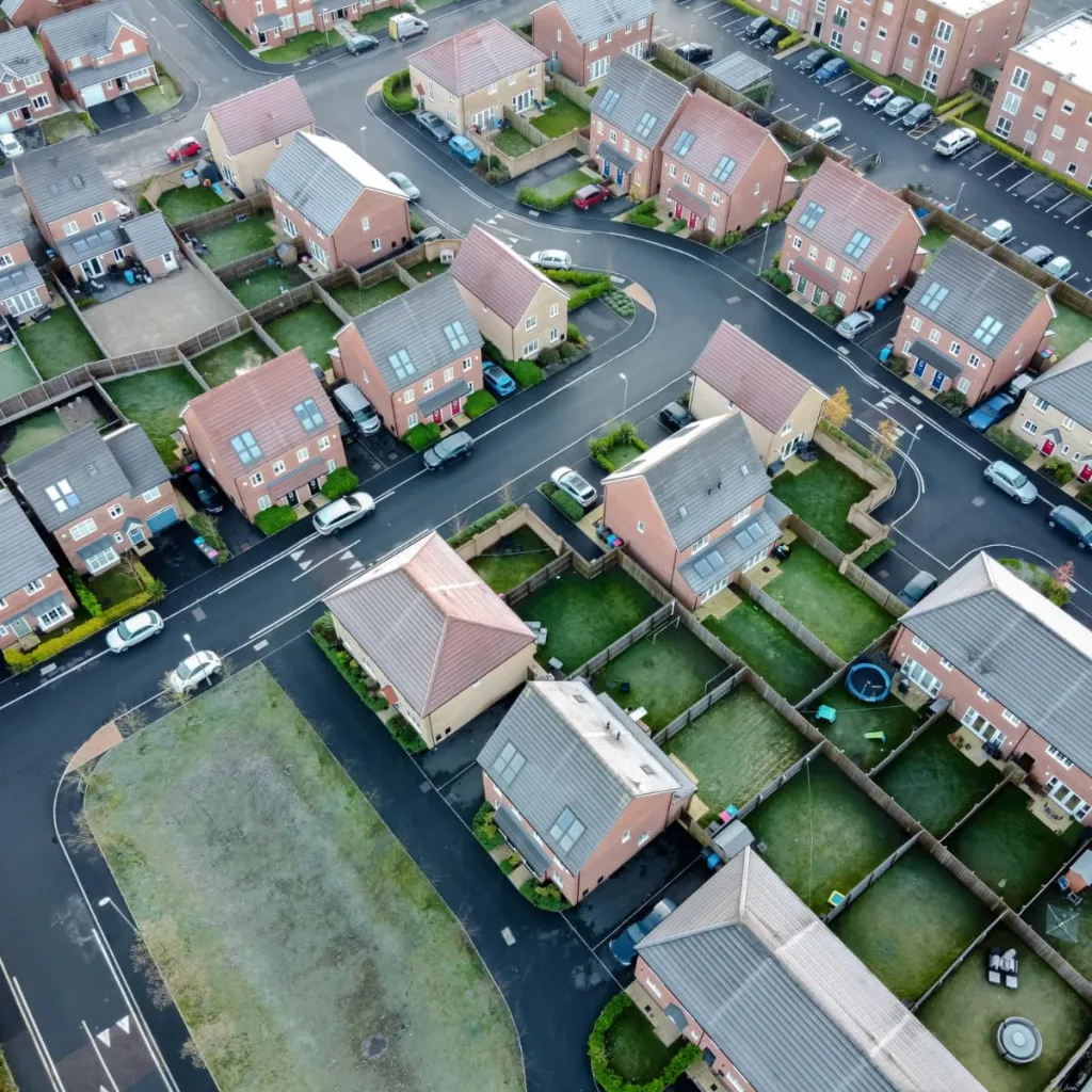Aerial view of a suburban residential street lined with identical red-brick semi-detached houses, each with small front gardens, driveways, and solar panels on their roofs, with parked cars along the street and green space visible on the left side.