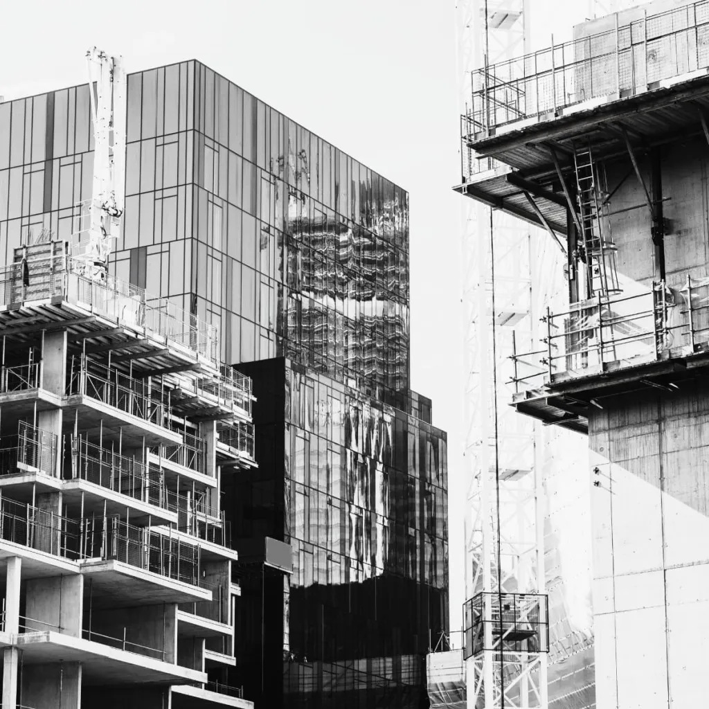 Black and white photograph of a modernist construction site with multiple high-rise buildings under development, featuring exposed concrete structures, metal scaffolding, and framework against a clear sky.
