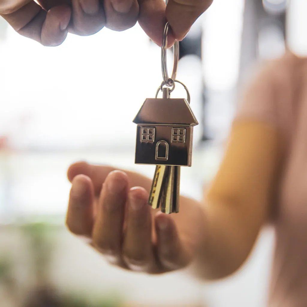A person's hand holds a house-shaped brass keychain with multiple keys attached, symbolizing home ownership and real estate decisions.
