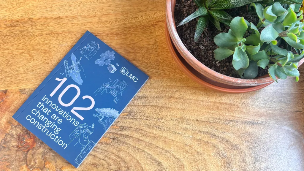 A blue hardcover book titled "102" with botanical illustrations sits on a wooden table next to a terracotta pot containing green succulent plants.