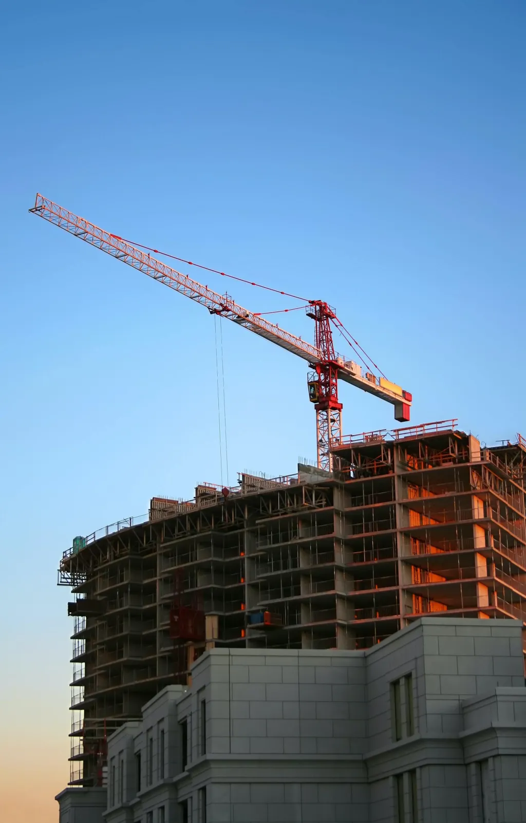 A large red and yellow construction crane extends diagonally across a multi-story residential building under construction against a clear blue sky, with a finished white stone structure visible in the foreground.