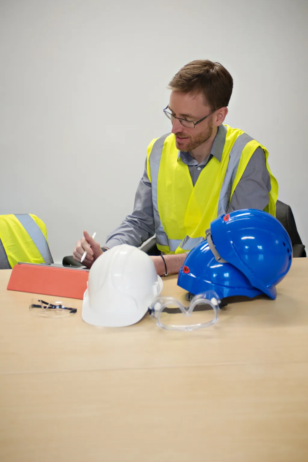 A man wearing glasses, a gray shirt, and a yellow safety vest sits at a table examining a blue hard hat while other safety equipment including a white hard hat and orange clipboard are visible on the table.