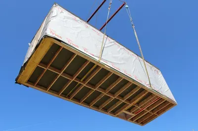 A large wooden structural beam or floor panel wrapped in white protective covering is suspended by cables against a clear blue sky, demonstrating prefabricated construction methods used in modern building projects.