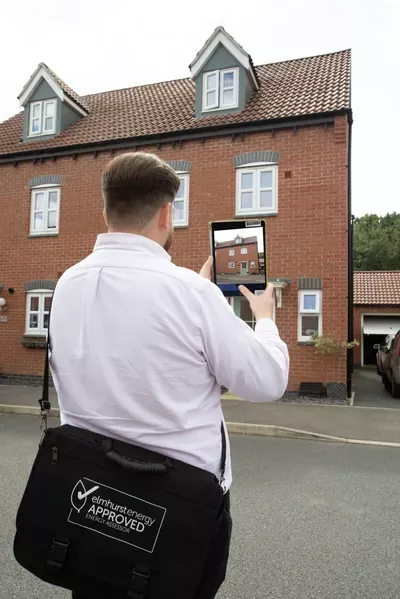 A surveyor in a white shirt and black apron holds a tablet while photographing the front facade of a brick residential house with dormer windows, likely conducting a property assessment or survey.