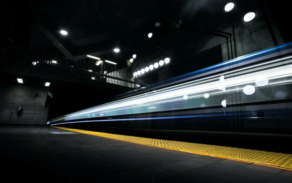 A subway train passes through a station platform at night, captured with motion blur to convey speed and movement, with blue and white lights trailing along the train's side and yellow tactile warning strips visible on the platform edge.