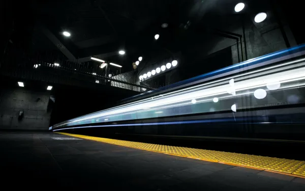 A subway train passes through a station platform at night, captured with motion blur to convey speed and movement, with blue and white lights trailing along the train's side and yellow tactile warning strips visible on the platform edge.