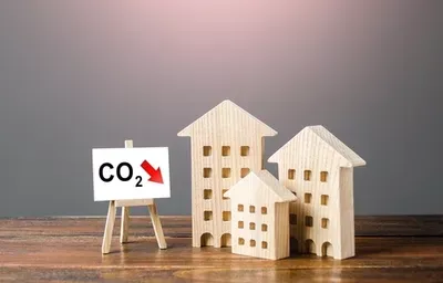 Wooden model buildings on a wooden surface with a small easel displaying a CO2 downward arrow on a gray background, representing sustainable construction practices.