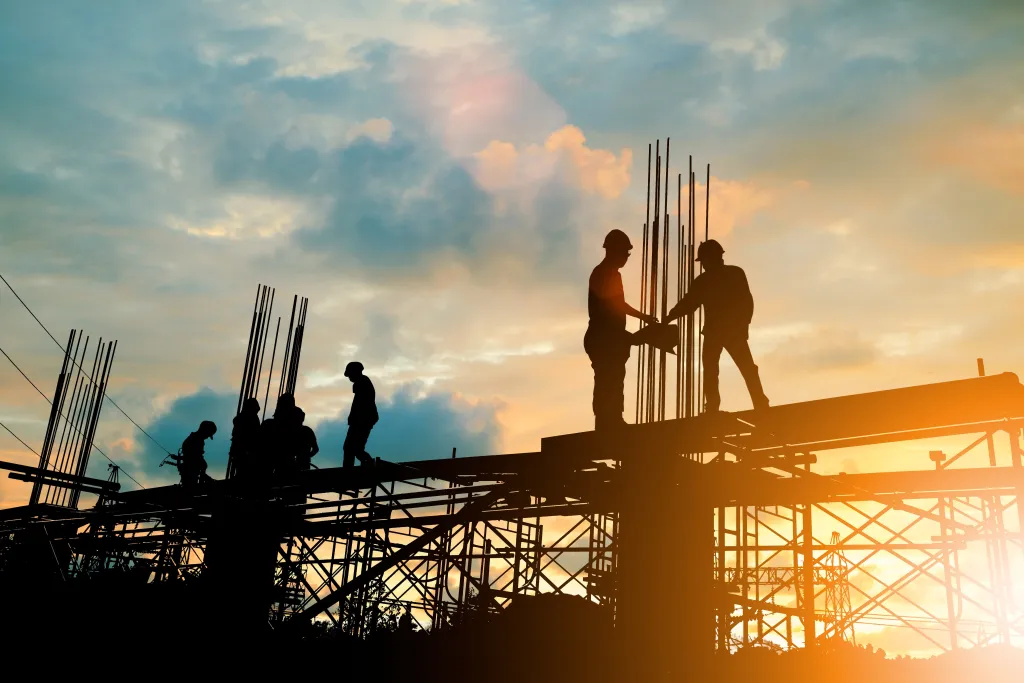 Construction workers silhouetted against a golden sunset, working on a high-rise building framework with exposed steel reinforcement rods and scaffolding visible across the structure.
