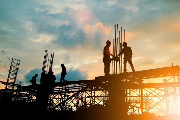 Construction workers silhouetted against a golden sunset, working on a high-rise building framework with exposed steel reinforcement rods and scaffolding visible across the structure.