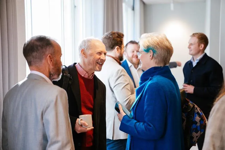 Thomas Lane at Building magazine greets Liz Male, founder of LMC, at Building magazine's roundtable debate on built environment education. Stood next to Thomas is Peter Barker from Ryder Architecture. In the background, a group of other participants are chatting.