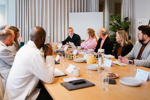 A group of men and women sit around a meeting room table, looking totwards a young man who is one of the PlanBEE programme graduates. This is a photograph taken at Building magazine's roundtable debate on built environment education.