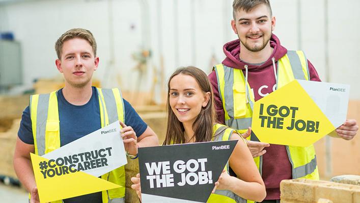 Three young people in high vis jackets from the PlanBEE programme hold up placards saying "Construct your career", "We got the job" and "I got the job".