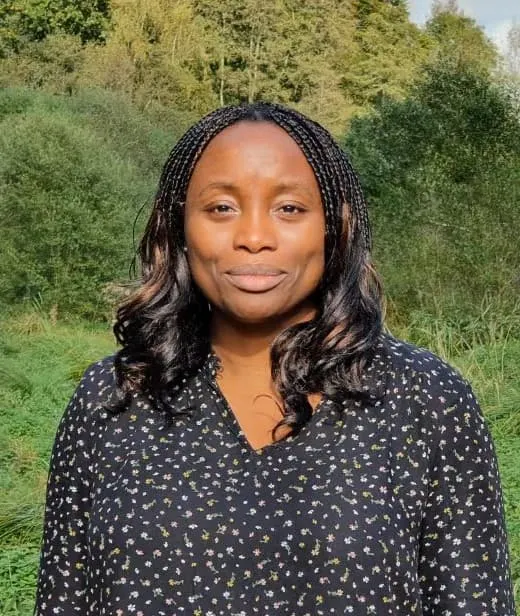 A woman with long dark braided hair wearing a dark patterned blouse stands outdoors with green foliage in the background.