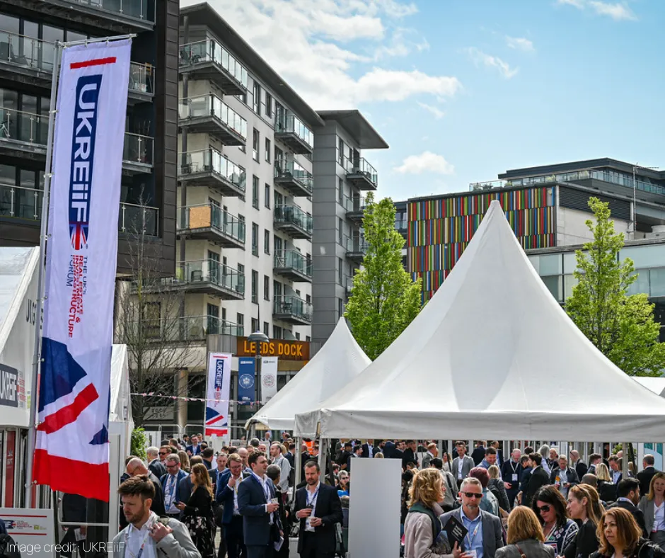 A large white tent structure hosts a crowded outdoor event in an urban setting, with multi-story residential and commercial buildings visible in the background. Attendees in business attire gather beneath the tent, with a blue and red banner and street signage visible to the left.