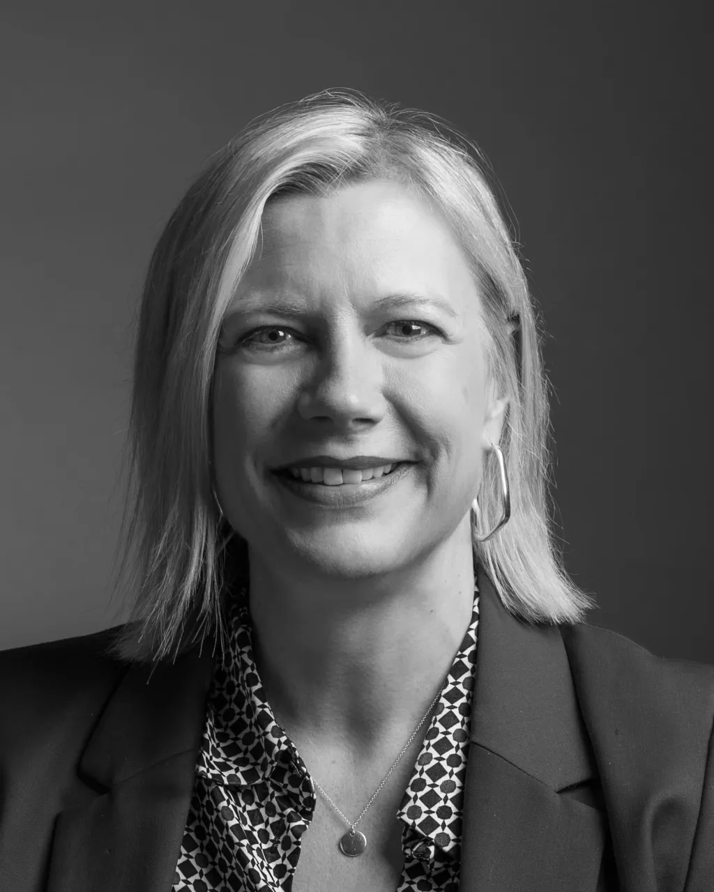 Professional headshot of a smiling woman with shoulder-length blonde hair wearing a dark blazer and patterned blouse with hoop earrings, photographed against a gray background.