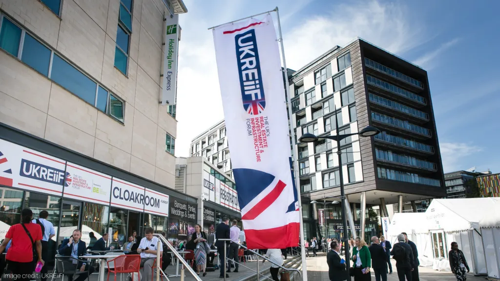 A busy outdoor event venue with modern buildings, featuring a large vertical UKREIF banner and a red-and-white striped flag, with attendees gathered around exhibition tents under a partly cloudy sky.